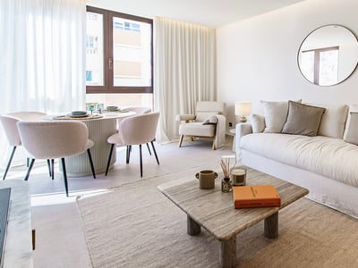 Salón luminoso con gran sofá blanco, mesa de piedra, espejos redondos y comedor junto a un ventanal.