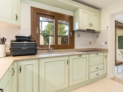 Ljust kök med mintgröna skåp, träfönster och mönstrat golv som leder till en matplats med korgstolar.