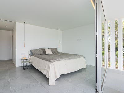 Minimalist bedroom with grey stone floors, white walls, and a large sliding glass door opening to a garden.