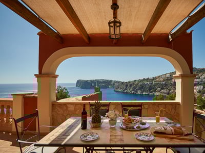 Terraza cubierta con mesa servida para el desayuno frente a una bahía azul y acantilados costeros bajo un cielo despejado.