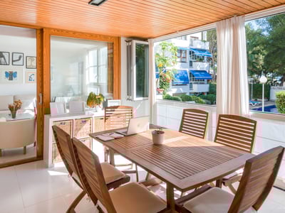 Geschlossene Holzterrasse mit Esstisch und Laptop, mit Blick auf Pool und Garten durch große Fenster.