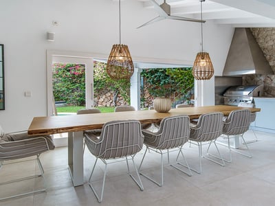 Mesa de madera maciza con sillas modernas y lámparas de mimbre junto a una cocina exterior con barbacoa de acero.