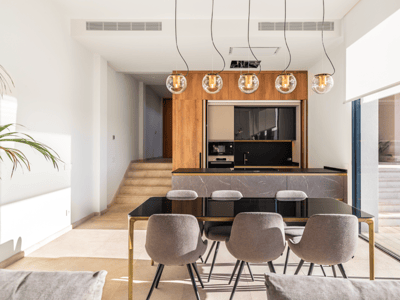 Comedor moderno con mesa de cristal negro, sillas grises, lámparas de diseño y cocina de mármol al fondo.