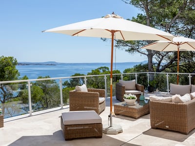 Sonnige Terrasse mit Loungemöbeln aus Korbgeflecht, weißen Sonnenschirmen und Glasgeländern mit Blick auf das blaue Meer.