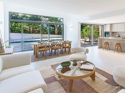 Bright open-plan room with modern white sofas, wood dining set, and minimalist kitchen overlooking a pool and garden.