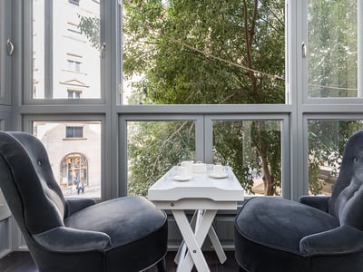 Zwei graue Samtsessel vor einem großen Fenster mit einem weißen Teetisch und Blick auf Bäume.