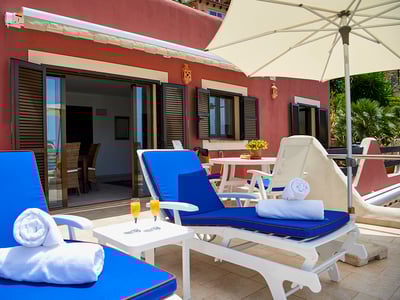 Terraza privada con tumbonas azules, toallas blancas, zumo de naranja y sombrilla frente a una casa color terracota.