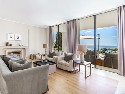 Modern living room with grey sofas, a stone fireplace, and floor-to-ceiling windows opening to a sea-view terrace.