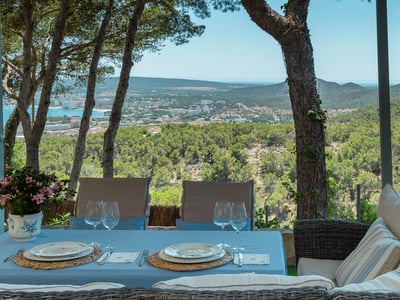 Outdoor dining table and wicker sofa on a shaded terrace overlooking a coastal town and sea.