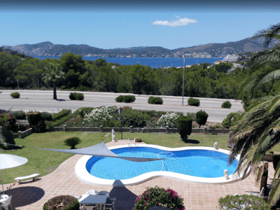 En oval pool i en trädgård med palmer, med utsikt över en blå vik och avlägsna berg.