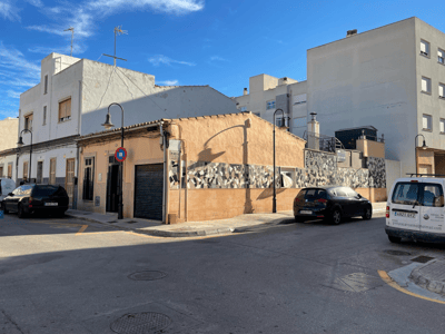 Pfirsichfarbenes Eckgebäude mit Mosaikstreifen in einer sonnigen Stadtstraße mit parkenden Autos.