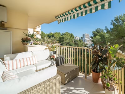 Terraza con sofás de mimbre blanco, plantas verdes y toldo a rayas con vistas a árboles y edificios.
