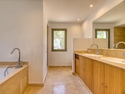 Baño de lujo con tocador doble de madera, encimeras de mármol beige, bañera y luz natural.