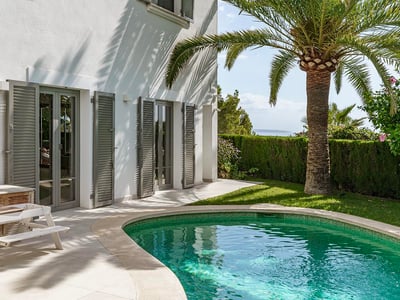 Piscina turquesa junto a una villa blanca con una palmera, persianas grises y vistas lejanas al mar.
