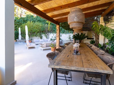 Mesa de comedor de madera bajo pérgola con sillas de mimbre y lámpara junto a salón exterior.