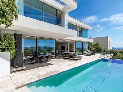 Villa blanca moderna con piscina, muebles de terraza y vistas panorámicas al mar bajo un cielo despejado.