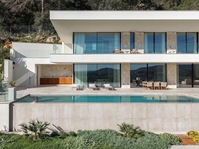 Modern two-story villa with a turquoise infinity pool, stone walls, large glass windows, and a sunny terrace.