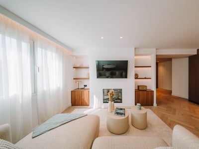 Ljust vardagsrum med vägg-TV, eldstad, fiskbensparkett och beige soffor.