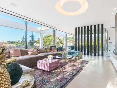 Modernes Wohnzimmer mit großem taupefarbenem Sofa, bunt gemustertem Teppich und bodentiefen Fenstern mit Blick ins Grüne.