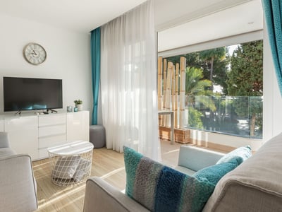 Modernt vardagsrum med grå soffor, turkosa kuddar och stora glasdörrar som leder ut till en lummig terrass.