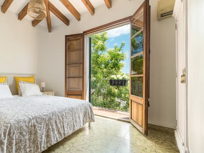 Un dormitorio luminoso con vigas de madera vista y puertas francesas que dan a un balcón soleado con árboles.