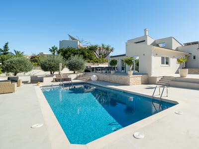 Klarblå pool intill en modern vit villa med stor stenterrass under en klarblå himmel.