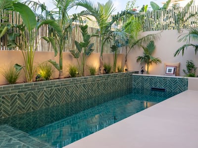 Lyxig pool med gröna fiskbenskakel, palmer och beige väggar med varm punktbelysning.