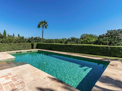 Piscina rectangular de azulejos turquesa rodeada de piedra natural y setos verdes bajo un cielo azul despejado.