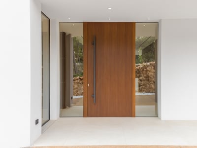 Large wooden pivot door with glass side panels flanked by smooth white walls and minimalist lighting.