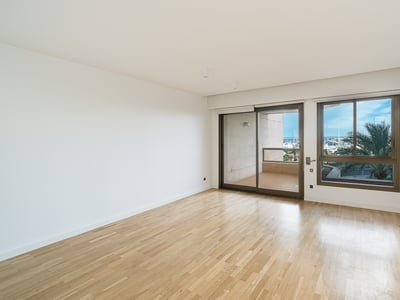 Habitación moderna vacía con suelos de madera clara y grandes ventanales con vistas a un balcón y un puerto deportivo.