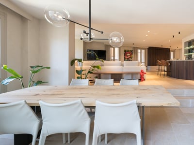 Großzügiger offener Wohnraum mit Esstisch aus Holz, weißen Stühlen, Glaspendelleuchten und einem Barbereich im Hintergrund.