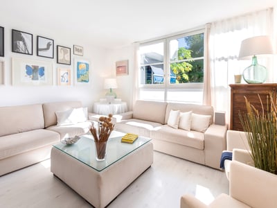 Helles Wohnzimmer mit zwei beigen Sofas, einem Glastisch, großen Fenstern und einer Wand mit gerahmter Kunst.