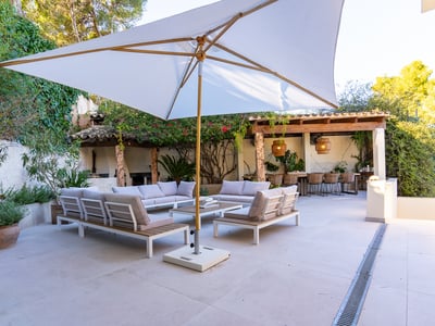 Amplia terraza con sofás modernos, sombrilla blanca grande y zona de comedor bajo una pérgola rodeada de vegetación.