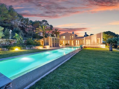 Longue piscine illuminée menant à une villa moderne en pierre sous un ciel de coucher de soleil éclatant.