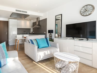 Ljust vardagsrum med grå fåtölj, turkosa kuddar, TV-bänk och modernt kök i bakgrunden.