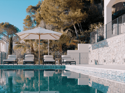 Infinity swimming pool with four sunbeds under a white umbrella, surrounded by stone walls and Mediterranean greenery.