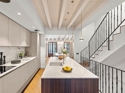 Cocina contemporánea con isla de mármol, armarios beige, vigas de madera y escalera de hierro negro.