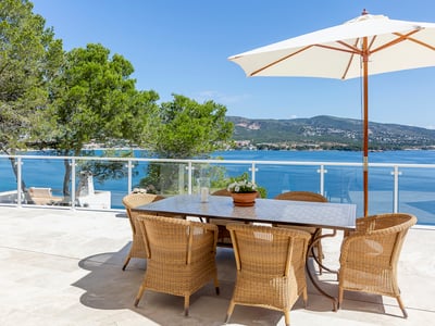 Außen-Essbereich mit Korbstühlen und weißem Sonnenschirm auf Steinterrasse mit Blick auf das blaue Meer und Berge.