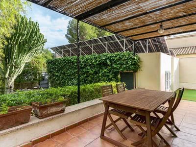 Matbord i trä under en bambupergola på en terrass med terrakottaplattor omgiven av växter och solpaneler.