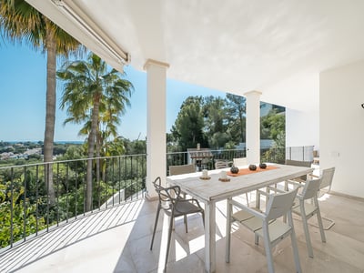 Vit terrass med matbord i trä, moderna stolar och utsikt över palmer och havet.