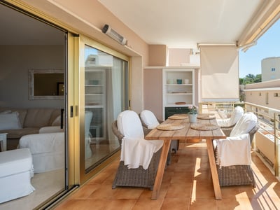 Terraza soleada con mesa de madera, sillas de mimbre y barandilla con vistas urbanas.