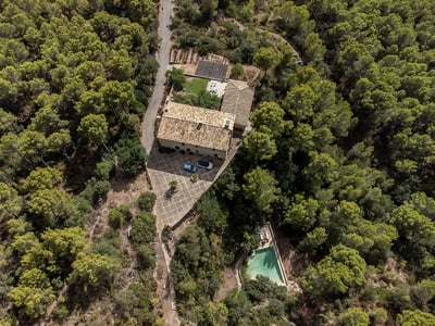 Aerial view of a stone house with terracotta roof, courtyard, and a pool, all surrounded by a dense green pine forest.