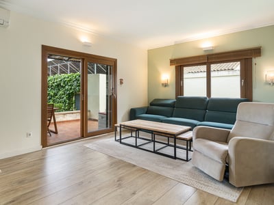 Ljust vardagsrum med blågrön soffa, fåtölj, trägolv och glasdörrar som leder till en lummig terrass.