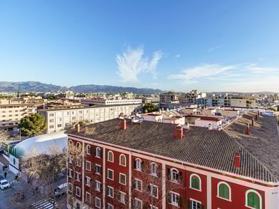 Vista aérea de una ciudad con un edificio rojo en primer plano, calles y montañas al fondo bajo un cielo azul.