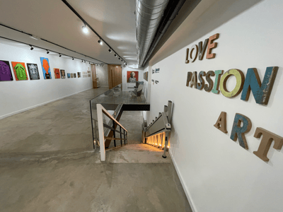 Galería de arte moderna con pinturas coloridas, suelos de cemento, una escalera de cristal y un elevador para sillas de ruedas.