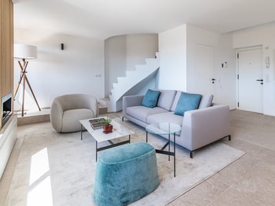 Sala de estar minimalista con sofá gris, otomana turquesa, escalera blanca y mesa de mármol.