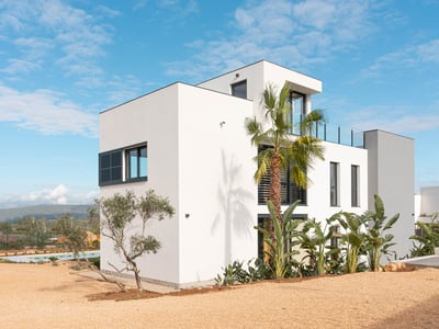 Exterior de una villa blanca de tres niveles con arquitectura moderna, palmeras y un patio de grava bajo un cielo azul.