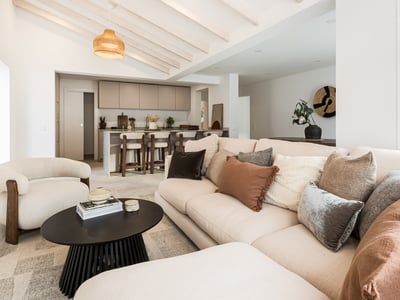 Salon lumineux avec canapé beige, poutres en bois et îlot de cuisine au second plan.