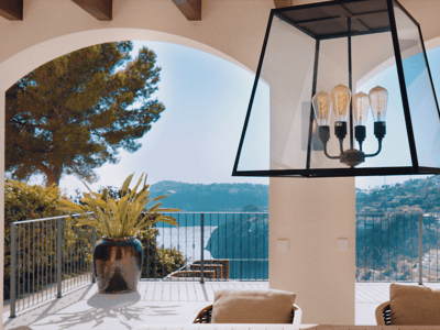View through white arches from a villa terrace overlooking the sea and green hills under a bright blue sky.