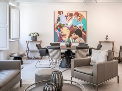 Habitación moderna y luminosa con un sillón de cuero gris, una mesa de comedor redonda y un gran cuadro colorido en la pared.